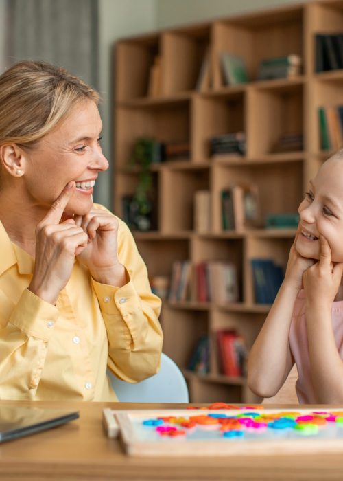 Female speech-language pathologist having lesson with little girl, teacher and pupil working on pronunciation, touching faces and grimacing, sitting at desk